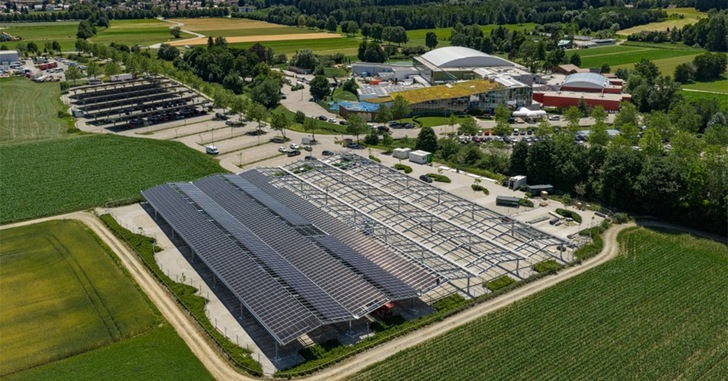 Luftaufnahme eines großen, mit Solarzellen bedeckten Parkplatzes in der Nähe von Feldern und mehreren Gebäuden. - © Timeless Planet GmbH & Co.KG