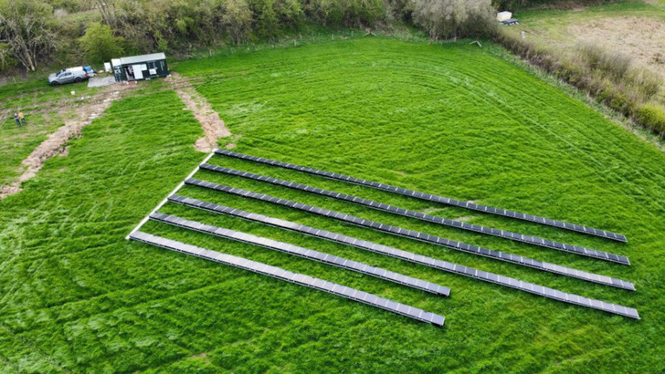 Die Solaranlage steht auf einer Wiese hinter einer Baumreihe. Daneben steht der Container mit dem Speicher und der Leistungselektronik.