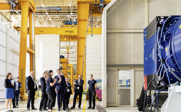 Führung einer Politikdelegation des Ministerpräsidenten von Nordrhein-Westfalen, Hendrik Wüst (Mitte, mit Mikrophon), durchs Winergy-Getriebewerk. Der Flender-Chef Andreas Evertz (Zweiter von rechts) lässt die Hochleistungsgetriebe vorzeigen.