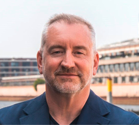 Lächelnder Mann mit kurzen grauen Haaren und Bart, der einen dunklen Blazer trägt, steht im Freien in der Nähe von Wasser und Gebäuden. - © Foto: Deutsche Windtechnik