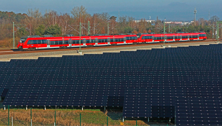 An einem klaren Tag fährt ein roter Personenzug hinter einem großen Feld von Sonnenkollektoren vorbei. - © Foto: Deutsche Bahn AG - Claus Weber