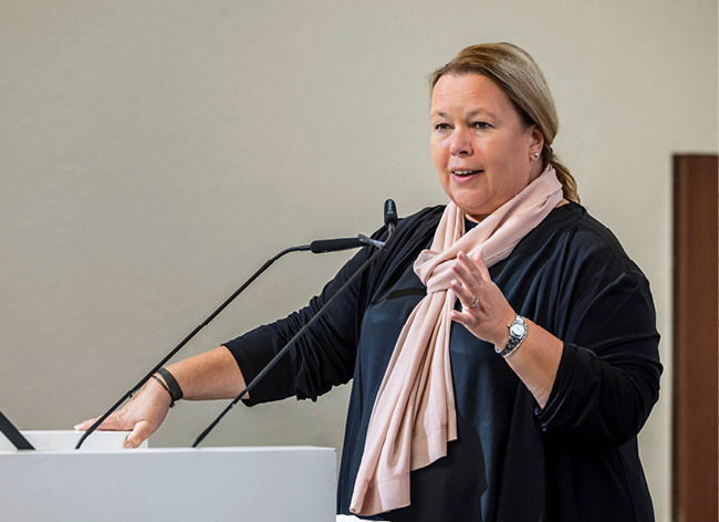 Eine Frau mit schwarzem Oberteil und rosa Schal spricht an einem Podium und gestikuliert mit einer Hand. - © Foto: BEE / Rolf Schulten