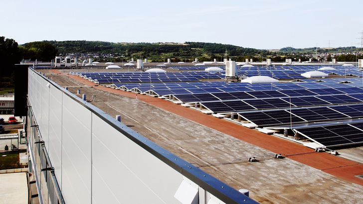 Reihen von Sonnenkollektoren auf einem großen Flachdach mit Bäumen und Hügeln im Hintergrund. - © BSW-Solar