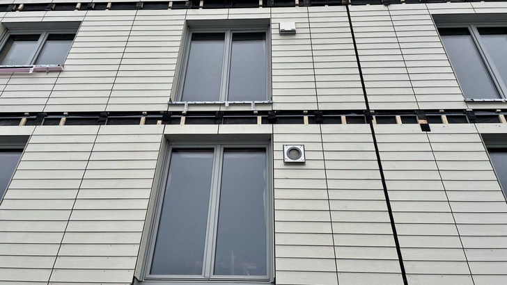 Nahaufnahme einer modernen Gebäudefassade mit grauer Verkleidung, drei Fenstern und einer Lüftungsanlage. - © dena