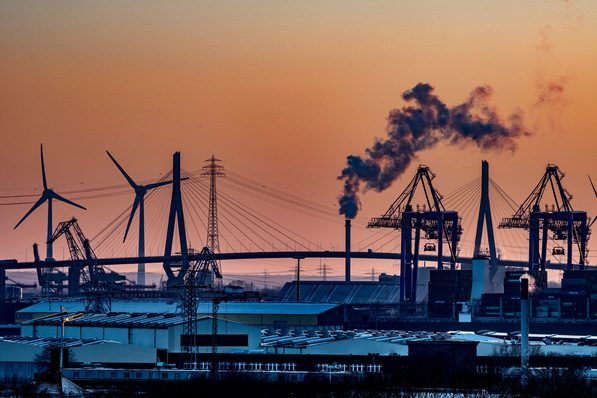 Industrielle Skyline bei Sonnenuntergang mit Kränen, Windturbinen und aus einem Schornstein aufsteigendem Rauch vor einem orangefarbenen Himmel. - © yourpix - stock.adobe.com