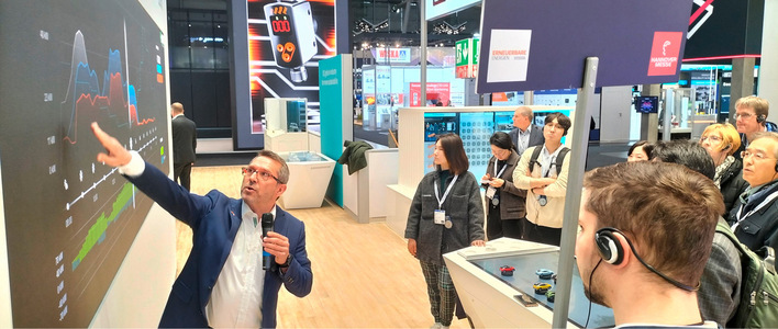 Ein Moderator erklärt einer Gruppe von Menschen auf einer Technologieausstellung Daten auf einem großen Bildschirm. - © Foto: Tilman Weber