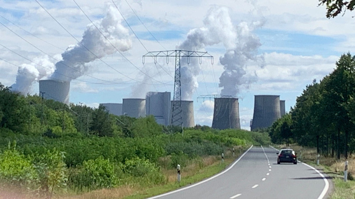 Eine Straße führt zu einem Kraftwerk mit dampfenden Kühltürmen, umgeben von Grün unter einem wolkigen Himmel. - © Nicole Weinhold