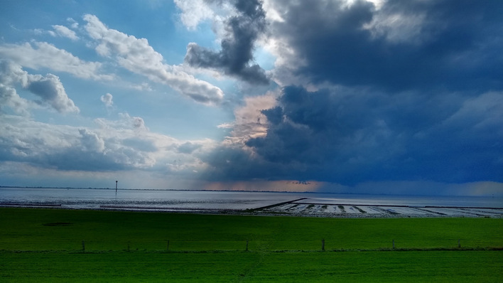 Dramatischer Himmel über einer weiten Landschaft mit grünen Feldern, Wasser und fernem Horizont unter großen, dunklen Wolken. - © Katharina Wolf