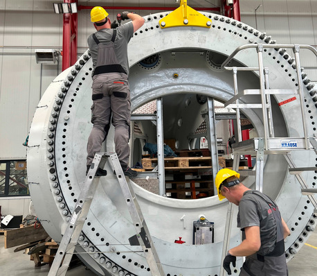 Zwei Arbeiter mit Schutzhelmen montieren oder prüfen in einer Fabrik mithilfe von Leitern ein großes Industriemaschinenteil. - © Foto: ENO ENERGY
