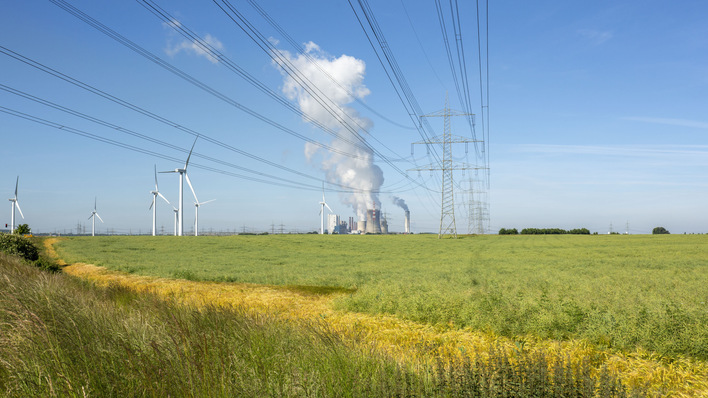 Windräder und Stromleitungen auf einer grünen Wiese, im Hintergrund eine rauchende Fabrik unter blauem Himmel. - © RWE