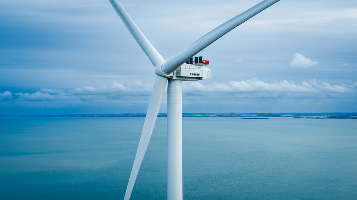 Unter einem bewölkten blauen Himmel steht eine große weiße Windturbine über dem Meer. - © Vestas