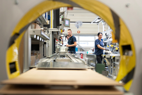 Zwei Personen bei der Arbeit in einer Fabrik, gesehen durch ein großes gelbes rundes Maschinenteil. - © Socomec